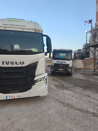 Two trucks are parked on a dirt and gravel lot in what appears to be an industrial area, possibly Polígon Industrial Dels Vents, Spain, based on the vehicle's license plates. The truck in the foreground, a white Iveco, has a license plate reading "7314 LKJ". The truck in the background, a white Mercedes-Benz with the company name "HORMAVAL" visible on its side, has a license plate reading "0107 KWX". Both trucks are facing forward, and the background shows industrial structures and scaffolding. The sky is clear and blue. The lighting suggests it is either early morning or late afternoon.