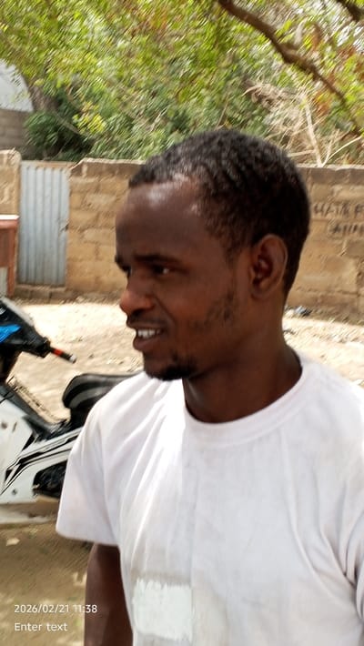 The image displays an outdoor scene in Hotoro South, Nigeria, dated 2026/02/21 at 11:38. A single adult male, with dark skin, short dark hair, and a short beard, occupies the foreground, positioned in profile facing right. He wears a plain white short-sleeved t-shirt, and his mouth is slightly open, revealing teeth. Behind the man, on the left, a portion of a white and black motorcycle or scooter is visible. The background features a light-colored masonry wall, a corrugated metal gate on the left, and dense green foliage from trees overhead. A white satellite dish is partially visible above the gate. The ground surface appears to be dirt or pavement.