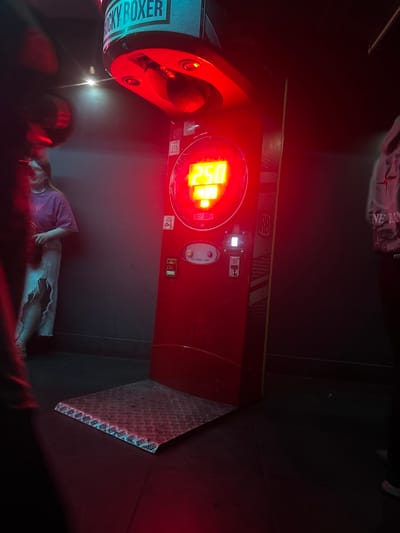 The image depicts a red "Rocky Boxer" arcade game under red lighting, displaying the score "250" on its illuminated screen. A metal platform is positioned in front of the game. To the left of the game, a young woman wearing a pink t-shirt and a light-colored skirt stands near a dark grey wall. To the right of the game, a person in a grey hooded jacket is partially visible. The scene appears to be an indoor entertainment venue, possibly an arcade or club, in Kirov, Russia, suggested by the "Rocky Boxer" machine, a popular type of game found in such establishments. The overall atmosphere is dimly lit with the dominant red glow from the game.