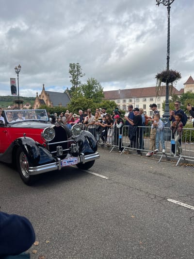 The image depicts a public event, likely the Bugatti Festival, in Molsheim, France. A vintage two-tone convertible automobile, predominantly black with a red body section, moves from left to right on an asphalt road. The car features prominent chrome components, including large circular headlights and bumpers, and displays a special event placard on its front bumper identifying it with "BUGATTI FESTIVAL MOLSHEIM" and the number "1." Multiple occupants, including a male driver wearing glasses, are visible within the vehicle.

On the right side of the road, a crowd of spectators of varying ages, including adults and children, is gathered behind silver metal crowd control barriers. Many individuals in the crowd are holding smartphones to record the event, and some adults are seen clapping. Children in the front row are observed standing on the lower bar of the barriers.

The background reveals several distinct architectural features of Molsheim. To the left-midground, an ornate, gothic-style stone chapel with a reddish-brown tiled roof is visible, partially obscured by green foliage. This building strongly resembles the Chartreuse de Molsheim. To the right-midground, a multi-story building with a reddish-brown tiled roof and light-colored facade is present. Further to the right, a structure featuring a turret-like element is visible, indicative of historic town architecture. Distant vineyards are visible on a hillside to the far left. The scene is illuminated by diffused natural light under an overcast sky. Two ornate black lampposts are present, one on the left displaying a banner that reads "BUGATTI FESTIVAL MOLSHEIM," and another on the right adorned with a hanging plant basket.