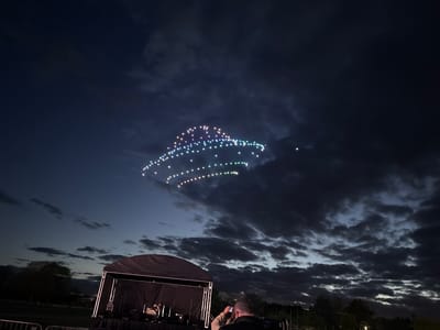 A nighttime scene in South Norfolk, United Kingdom, showing a stage with equipment in the foreground. Behind the stage, a person holds up a mobile phone, potentially photographing or recording the scene. The sky is dark with heavy clouds. In the upper part of the frame, an illuminated aerial display of lights in the shape of a rounded object is suspended against the night sky. Trees are visible in the background.