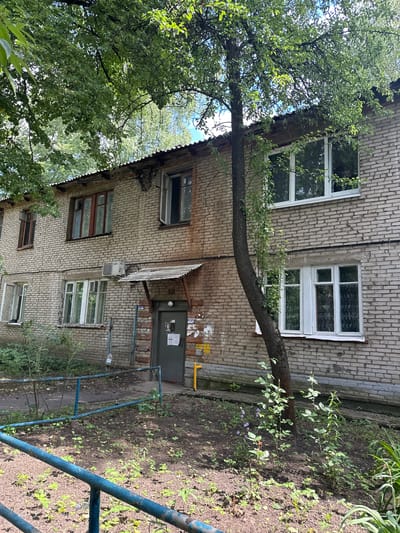 The image shows a two-story brick apartment building in Lytkarino, Russia, with a tree growing near the front of the building. The building's facade is constructed of light-colored bricks. It has a corrugated metal roof. Several windows are visible, some with white frames and others with reddish-brown frames. One window on the upper floor is open. An air conditioning unit is mounted below a window on the second floor. A small, weathered awning extends over a doorway on the ground floor. The ground in front of the building is covered with dry earth and sparse vegetation, with a rusty blue metal railing in the foreground. The overall scene appears to be an outdoor residential area.