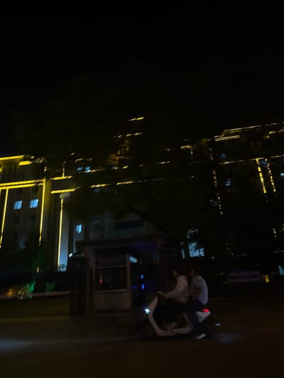 At night, in Jinzhou, China, a view from a window reveals city lights and building facades illuminated with linear yellow lights. Two individuals are visible; one standing and another lying down. The lighting appears dim, and the image lacks sharpness.