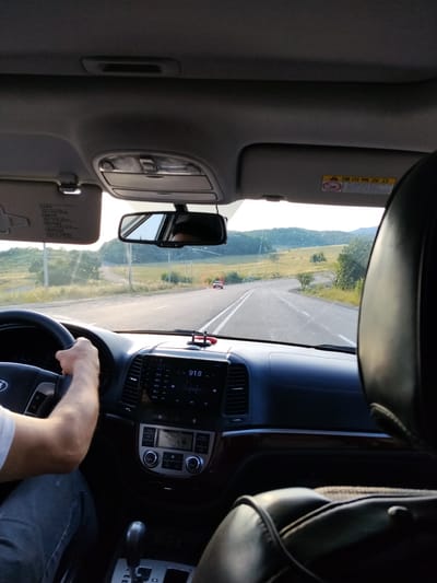 The image shows the interior of a car from the perspective of a passenger. A person's hands are visible on the steering wheel, indicating they are driving. The dashboard is prominent, with a touchscreen display showing "91.8" and other radio frequencies. To the right, a dark leather headrest of a seat is visible. The car is driving on a paved road that winds through a hilly, rural landscape with green fields and scattered trees. A red car is visible further down the road. The sun is low in the sky, casting long shadows and creating a warm, golden light, suggesting it is either early morning or late afternoon. The sky is clear. There is text visible on a yellow warning sticker on the sun visor, which appears to be in Korean. The scene suggests a leisurely drive through the countryside in the Ismailli District of Azerbaijan.