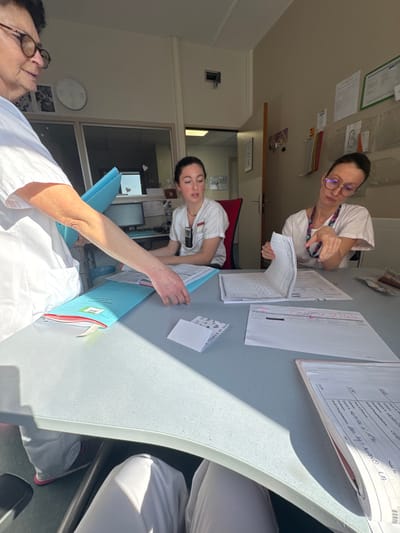 An indoor scene in a healthcare facility in Tourcoing, France, depicts three adult females in medical uniforms engaged in administrative tasks around a light gray, curved table.
On the left, a partially visible older female, wearing glasses and a white uniform, uses her right hand to place a blue folder containing documents onto the table. Seated centrally is a younger female in a white scrub top with a clipped communication device, looking attentively towards the left. On the right, a middle-aged female, wearing glasses and a white scrub top with a lanyard, actively reviews paper documents, holding an open form with both hands.
The table is covered with various documents, including blue folders, loose papers with handwritten notes and grids, and a small white card partially labeled "ST VA" and "centre chance et soci." A strong diagonal shadow is cast across the table from an unseen light source. In the background, a desk with a computer monitor, an unoccupied red office chair, and a closed door are visible through a windowed partition.