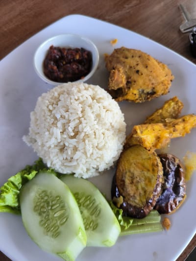 On a square white plate, a meal is arranged. A mound of white rice is centrally located. To the left, two cucumber slices lie on a bed of lettuce. Adjacent to the rice, a small white bowl contains a dark red sauce. To the right of the rice, there's a piece of fried chicken along with fried bananas, and fried eggplant. This scene likely takes place in Matang, Indonesia. The presence of a car key at the top-right corner suggests the plate is on a table, possibly in a restaurant or home setting.