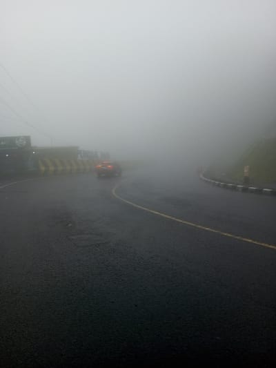 A car navigates a winding road obscured by heavy fog. Road markings, including a yellow line and black and white barriers, are partially visible. A building is present to the left of the road. Poor visibility characterizes the scene, suggesting hazardous driving conditions. The asphalt road surface appears wet.