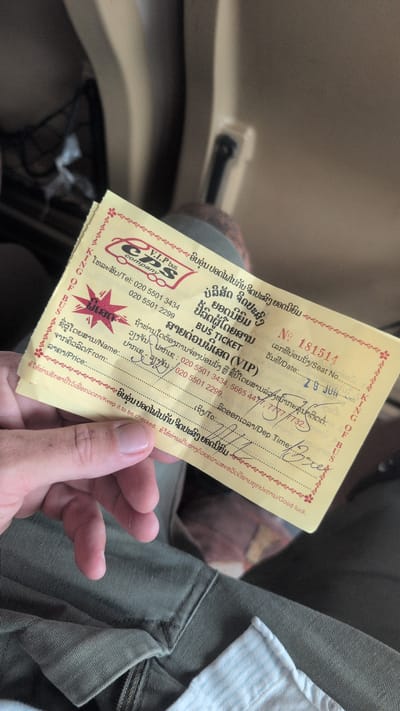 A bus ticket is held by a person's hand in an indoor environment, possibly a vehicle. The ticket is yellow with red and black text and graphics. The text is in both Lao and English. The Lao text appears to be for a bus company named "CPS V.I.P. bus company." The ticket has a serial number "No. 181514" and a date that appears to be "29 JUN". There are handwritten entries on the ticket, including a name that appears to be "Sorn" and a departure time and date. The background of the image is blurred, but it appears to be the interior of a vehicle with beige seating. The person's hand is holding the ticket with their thumb and index finger.
