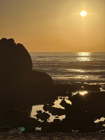 A sunset over the ocean is depicted, with the sun a bright orb in the upper right quadrant of the frame, casting a golden reflection on the water. The waves are gently rolling towards a rocky shore in the foreground. The rocks are dark and silhouetted against the bright water and sky. In the bottom left corner, there is a large, dark rock formation that dominates the left side of the image, its jagged edges outlined by the fading sunlight. The sky is a warm gradient of orange and yellow. The scene appears to be a natural coastal environment, possibly a beach or coastline. The image does not contain any specific landmarks that would definitively place it in Biarritz, France, but it is a generic coastal sunset scene. There are no people visible in the image.