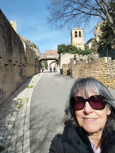 A woman with gray hair and large sunglasses poses in the foreground. Behind her, a narrow paved road stretches between high stone walls. An arched walkway spans the road in the distance, with figures walking beneath. A church tower rises to the right, while a tall, thin tower stands to the left. The scene is bathed in sunlight under a clear blue sky. Bare branches of trees are visible above the church tower.