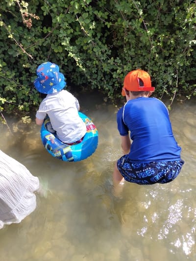 An outdoor scene depicts three individuals in shallow, clear, light-brown tinted water, positioned against a dense backdrop of green leafy foliage, located within the Maale Yosef Regional Council, Israel.
To the immediate left, the lower portion of an individual, clad in a light-colored, pleated garment, is visible, standing in the water.
In the center-left, a young child, viewed from the rear, wears a white long-sleeved shirt and a blue bucket hat patterned with white stars and red elements. This child is seated within a blue inflatable swim ring featuring a checkered pattern and various graphics, facing towards the foliage.
To the right, a second child, also viewed from the rear, wears a blue long-sleeved swim shirt, dark blue swim shorts with white text graphics, and an orange baseball cap. This child is standing in the water, leaning forward slightly.
The water's surface exhibits numerous sun reflections, and the bed beneath is stony. The background consists of a dense growth of green leaves and branches, extending from the water's edge upwards.