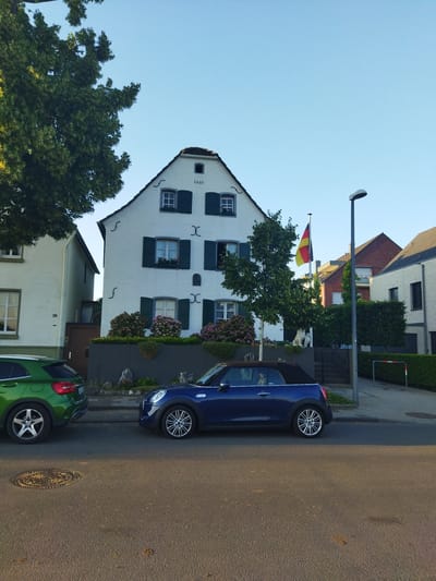 The image shows a street scene in Leverkusen, Germany, with a focus on two parked cars and a white, historic-looking building. A green SUV is parked on the left, and a blue convertible is parked on the right. The white building, which appears to be a house, has dark green shutters on its windows and a date "1661" inscribed above the top-floor windows. A German flag is visible on a flagpole in front of the building. The sky is clear and blue, suggesting it is daytime. The street is paved, and there is a sidewalk with a hedge and a street lamp. Other houses and trees are also visible in the background.