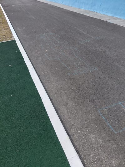 This media file presents an aerial-view image of an outdoor recreational area, likely a playground or sports court, located in Nadym, Russia. The scene is dominated by a vast expanse of dark gray asphalt pavement, showing a textured, granular surface.
To the left, a vibrant blue painted wall rises, bordered by a concrete coping that separates it from the asphalt. A stark white painted curb or dividing line runs diagonally across the lower right section of the image, segmenting the asphalt from a rich dark green, textured surface, which appears to be artificial turf or a rubberized safety surface. In the bottom left corner, a small section of dry, light brown grass and dirt is visible, suggesting a transition to natural ground.
The primary activity suggested in the image is children's play, although no individuals are present. This is evidenced by multiple drawings sketched with light blue chalk on the asphalt. The most prominent drawing is a complex, non-standard hopscotch-like grid featuring various interconnected boxes and lines. Within some of these boxes, faint and irregularly drawn letters or symbols can be discerned, such as what appears to be "D", "K", and "L", though their complete interpretation is challenging due to their faded nature. Another simpler, square-shaped chalk outline is visible further up and to the right on the asphalt.
Notable details include the bright, clear lighting, indicating it is daytime, likely sunny, given the sharp contrast and lack of long shadows. The overall impression is one of a quiet, prepared space awaiting or having recently hosted recreational activities.