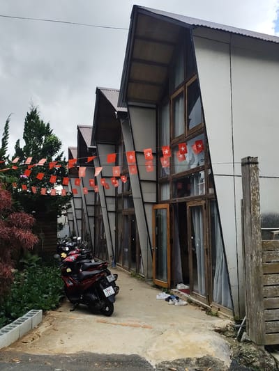 The image shows a row of triangular-shaped buildings with corrugated metal roofs and light gray siding, located in Lam Vien - Da Lat, Vietnam. The buildings have large wooden-framed windows and doors. A string of small orange flags with white stars is strung across the front of the buildings, hanging from a wire. In front of the buildings, a paved walkway is lined with dense green foliage and a large tree. Several motorcycles are parked on the walkway. A pair of white sneakers and a pair of blue and white sneakers are placed near the entrance of one of the buildings. The sky is overcast.