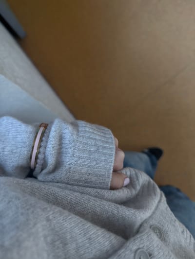 A top-down shot features a person's clothed arm, adorned with bracelets, resting against a light grey cardigan and blue jeans. A glimpse of a black shoe is visible. The cardigan has visible buttons. The bracelets are thin, one gold with a pink band and another string of light-colored beads. The hand partially obscured by the sleeve, with light pink nail polish visible. The setting appears to be indoors, with a tan wall in the background. A white surface is partially visible below the arm.