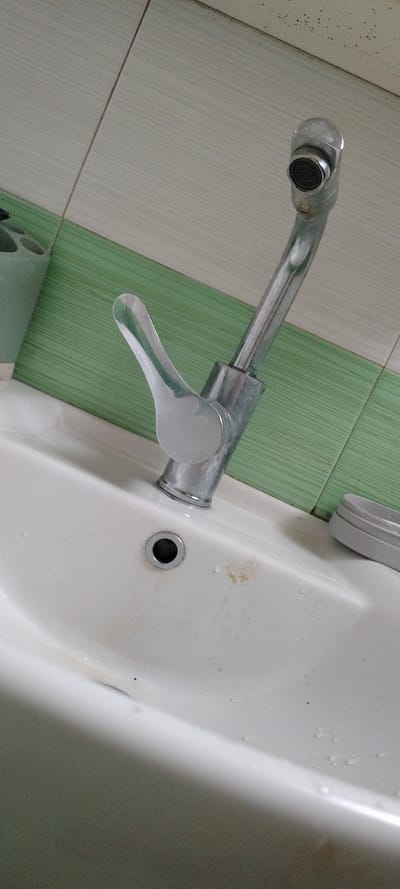 The image presents an indoor scene featuring a chrome-finished single-lever faucet positioned over a white ceramic sink. The faucet has a long, slender spout with a black aerator visible at its end, and a flat, reflective lever handle.

The white ceramic sink shows small water droplets and a light reddish-brown discoloration or stain near its circular metal drain. A portion of a grey, possibly plastic or metal, ring or cover is visible on the upper right edge of the sink.

The background features tiled walls with alternating horizontal stripes of light off-white and light green. The off-white tiles exhibit a subtle linear texture. No specific activity is occurring, and there is no visible text. The setting appears to be a bathroom or utility area in Beguntsi, Bulgaria. The image is captured from a slightly elevated, diagonal perspective.