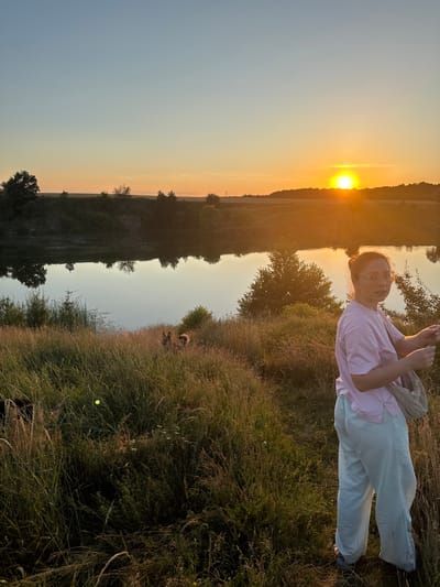 The image captures a young woman in a pink t-shirt and light blue pants standing in a grassy field during sunset. The sun is low on the horizon, casting a warm, golden glow across the sky and reflecting on a body of water. The woman is facing the camera with a slight smile, her hair pulled up in a bun, and she is wearing glasses. She has a light-colored bag slung over her shoulder. The foreground is filled with tall, dry grass, suggesting a natural, outdoor setting. In the background, there are trees and rolling hills. The overall atmosphere is peaceful and serene, with the warm light of the sunset creating a calming mood. The scene appears to be set in Horodyshche, Ukraine. There is no visible text in the image.