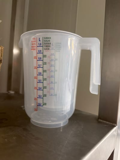 A clear plastic measuring cup with a handle sits on a stainless steel surface. The cup has multiple measurement markings on its side in different colors. The markings include "L" for liters, and scales for "MEL FLOUR," "MEHL," "MAKA," and "FARINE" in red text. There are also markings for "SUKKER," "SUGAR," "ZUCKER," "CUKIER," and "SUCRE" in green and blue text. The liquid level inside the cup appears to be at the 0.2 L mark. The background is a neutral, metallic wall. There is no indication of the city of Częstochowa, Poland in the image.