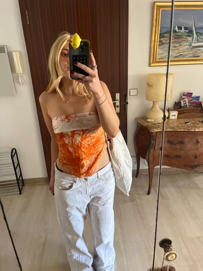 The photograph appears to be taken in an interior setting, likely a home. The primary subject is a young woman holding a smartphone, seemingly taking a selfie in front of a large mirror. Her top is patterned with orange, coral-like designs, and she is wearing white pants. She has blonde hair and jewelry on her fingers and wrist. The phone has a yellow charm attached to it.

The mirror reflects a scene that includes a wooden dresser, an ornate painting in a gold frame, and a lamp. There is also a pillow on the dresser. The floor is tiled in a light beige color. There is also a heating panel on the wall and a radiator rack, on the wall behind and beside the girl respectively.

Given the presence of the painting, dresser, and the architectural style reflected in the mirror, the location could be a private residence or possibly a hotel room. However, it is impossible to verify the exact location within the city of Antibes, France, with certainty without additional visual clues or contextual information.