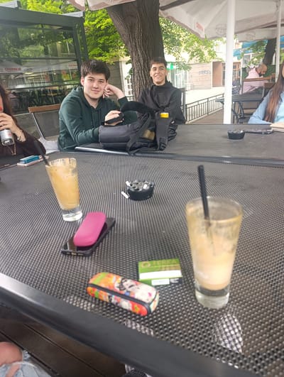 The image shows a table at an outdoor cafe, likely in Silistra, Bulgaria. Two young men are seated at the table, facing the camera. One is wearing a dark green hooded sweatshirt and has his head resting on his hand. The other is wearing a black hooded sweatshirt and is smiling. Between them is a black backpack. On the table are two glasses containing beverages, an ashtray with cigarette butts, a pink device on top of a smartphone, a multicolored case, and a pack of cigarettes. Other people are visible in the background seated at other tables. Trees and buildings are also visible.