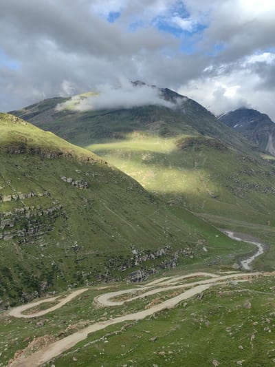 The image captures a scenic mountain landscape in Gramphu, India. In the foreground, a winding dirt road with several switchbacks carves its way up the steep, green mountainside. The road is bordered by rocks and sparse vegetation. The slopes are covered in lush green grass, with rocky outcrops and ledges visible. A thin, winding river flows through the valley in the middle ground. The background features towering mountains, partially obscured by dramatic, heavy clouds. Sunlight intermittently breaks through the clouds, casting bright patches on the mountain slopes. The sky is a mix of gray clouds and patches of blue. No people or vehicles are visible in the frame.