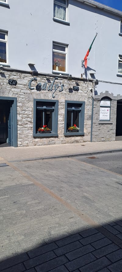 The image shows the exterior of a building with a stone facade. The building has several windows, some with flower boxes. A sign on the building reads "Coady's Funeral Home" and includes a website address "www.castlebarfuneralhome.ie". Another sign appears to say "Est 1905". The building has a white upper section and a grey stone lower section. A flag, possibly the Irish flag with an added green element, is mounted on the wall. The scene appears to be outdoors on a sunny day, with shadows cast on the pavement in front of the building. The pavement is made of stone slabs. The overall impression is of a historic building in a town.