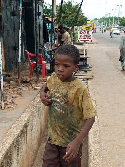 A boy stands on a dirt path next to a concrete retaining wall in Garko, Nigeria. The boy is wearing a yellow, stained t-shirt with a graphic design and dark brown shorts. He is looking to his right and appears to be holding something small in his right hand. To his left, there is a building with a dark metal door, a red chair, and a bicycle. In the background, a street with traffic, including cars, motorcycles, and a yellow truck, is visible. A sign reading "POS AND DATA" is also visible. The environment appears to be an urban setting with various buildings and infrastructure.