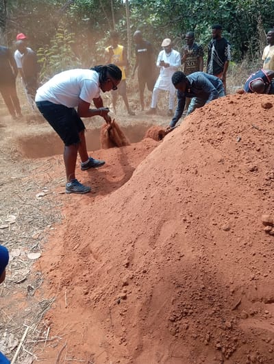 The image depicts an outdoor scene in Okpatu-Ukehe, Nigeria, characterized by dry, reddish-brown earth. Two men in the foreground are actively engaged in moving soil next to two excavated trenches. The man on the left, wearing a white t-shirt, dark shorts, sunglasses, and dreadlocks, is bent over, scooping reddish-brown soil into a light-colored sack from a mound adjacent to a trench. The man on the right, dressed in a dark patterned shirt and dark trousers, is also bent, moving soil from a larger pile with his hands or a small tool, pushing it towards or into an adjacent trench. Visible dust particles are suspended in the air. Approximately eight to ten additional men are standing in the midground and background, observing the activity. They are clad in varied casual attire, including t-shirts, a yellow basketball jersey with "22," a dark long-sleeved shirt, and a white traditional tunic and trousers. One observer in white is looking at a mobile phone. The ground consists of dry, reddish-brown soil with minimal dry vegetation. A dense cluster of green trees and bushes forms the background.