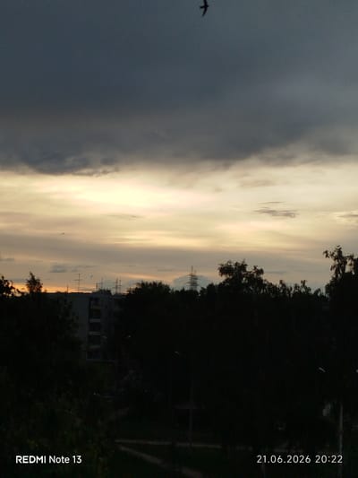 An overcast sky with a band of bright, possibly sunset light, stretches across the horizon. Dark silhouettes of trees and buildings are visible in the foreground, obscuring much of the urban landscape. The city of Stroykeramika, Russia, is implied by the presence of residential buildings and what appear to be power lines or transmission towers in the distance. A lone bird is captured in flight against the dark, upper portion of the sky. The date and time stamp "21.06.2026 20:22" and "REDMI Note 13" are imprinted on the lower right and left corners of the image, respectively.