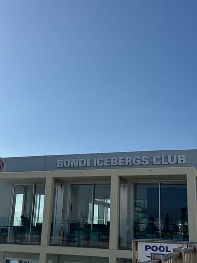 The Bondi Icebergs Club building is pictured against a clear blue sky. The vertical view highlights the club's name prominently displayed in large letters. Windows reflect the outdoor scenery. A sign directs towards the "POOL." The architectural design showcases clean lines and a modern aesthetic. The club's location is seemingly near the coast due to the reflective windows.