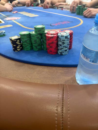 The image captures an angled, close-up view of a poker game in progress, set indoors. The primary focus is on a blue poker table laden with various stacks of poker chips and a water bottle.
**Subjects:** Several sets of hands belonging to players are visible around the table, engaged with chips and cards, but no faces or full figures are discernible.
**Objects:** Dominating the foreground and midground are numerous stacks of poker chips. These include prominent stacks of red, light blue, green, and black chips. The red and light blue chips clearly display "CASINO" along with other branding. Green chips have a lighter central design, and black chips feature yellow markings. The blue poker table is made of a felt-like material, marked with a yellow curved line and a faint red "CASINO" logo. A clear plastic water bottle, partially filled, rests on the table's edge. A few red-backed playing cards are visible on the table. The right side of the frame features a brown or tan, leather-textured surface, likely a chair armrest or table trim, characterized by horizontal stitching.
**Activity:** The presence of multiple players' hands, poker chips, and playing cards clearly indicates an active poker game.
**Notable details:** The setting is an indoor environment, suggested by artificial lighting. Textural variety is evident, from the smooth plastic of the bottle and the textured chips to the soft felt of the table and the stitched, textured leather-like material of the armrest. The overall atmosphere appears concentrated on the game. While the image itself provides no specific visual cues for its location, the provided context indicates it is in Larnaca, Cyprus.
**Visible text:** "CASINO" is legible on some red and light blue poker chips, and faintly on the blue table surface. The water bottle's label clearly shows "0.5L" along with a barcode and other smaller, unreadable text.
