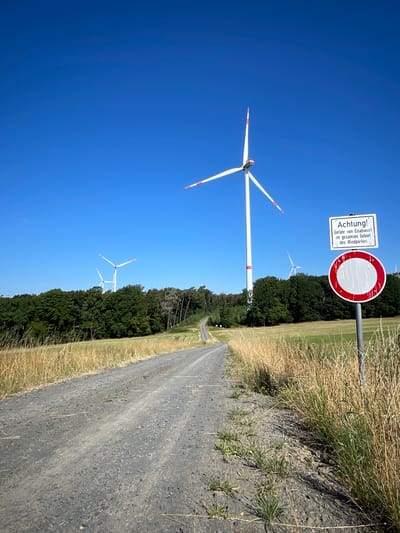 In the city of Berfa, Germany, a gravel road winds through a grassy field towards a wind farm under a clear blue sky. In the foreground, dry grass lines the side of the road. To the right, a signpost displays a warning message in German, reading "Achtung! Gefahr von Eisabwurf im gesamten Gebiet des Windparkes" (Attention! Danger of ice falling in the entire area of the wind farm), along with a round, red and white prohibitory sign. Several wind turbines are visible in the middle ground and background, their white towers and red-tipped blades stark against the sky. The turbines are positioned amidst a line of dense green trees. The scene depicts a rural landscape dominated by renewable energy infrastructure.