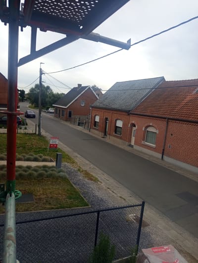 The image shows a street scene in Lebbeke, Belgium. It appears to be daytime with overcast skies, suggesting a cloudy or potentially rainy day. The perspective is from an elevated position, looking down a residential street lined with brick houses.
In the foreground, there are elements of construction scaffolding, including red and silver metal poles. A black chain-link fence is also visible, with a patch of gravel and some low-lying grass or ornamental plants beyond it. A "TE KOOP" (For Sale) sign is planted in the grass, indicating that at least one of the properties is on the market. The sign has text including "Grond + woning" (Land + house) and a phone number.
Further down the street, several cars are parked or driving. The houses themselves are typical of a European residential area, with red tile roofs and brick facades. Some houses have visible windows, some with blinds partially drawn. A street lamp is visible in the distance, along with power lines and trees. The overall atmosphere is quiet and suburban. There are no people clearly visible in the scene, but the presence of a "For Sale" sign suggests an ongoing real estate transaction.
