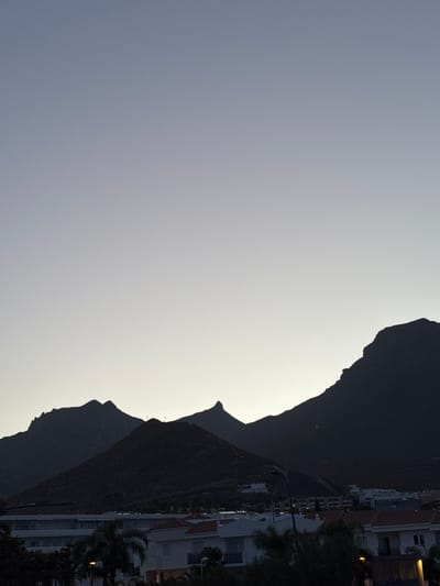 This is a vertical, medium-shot photograph of a mountainous landscape above a town at dusk, taken from a rooftop. The sky is a gradient of pale grey and light blue, with the palest color at the horizon. Silhouetted against the sky are three dark, jagged mountain peaks. The mountains are covered in scrub and vegetation, and a faint road or path can be seen winding up the side of the central mountain. Below the mountains is a town with white buildings and red-tiled roofs. The buildings appear to be hotels or apartment complexes, with balconies and windows visible. Palm trees and other foliage are scattered throughout the town. A lamppost stands to the left of the frame. The overall mood is serene and peaceful, with a sense of quietude as night approaches. The scene is consistent with the landscape of Adeje, Spain, known for its volcanic mountains and coastal resorts.