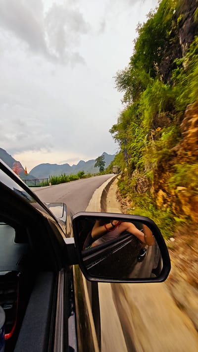 The image depicts a car traveling on a road in Cán Tỷ Commune, Vietnam. The view is captured from inside the car, looking out through the side window and side mirror. In the background, there are mountains and a cloudy sky. The road curves along a hillside, with green vegetation visible on the side. The side mirror reflects a person with their arm out the window. The car appears to be in motion, as evidenced by the blurred background.