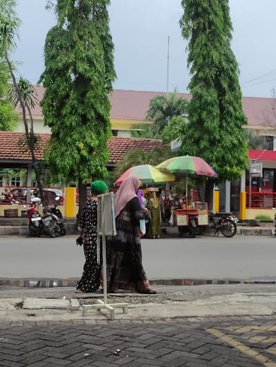 This is a daytime, eye-level shot of a bustling street scene in Situbondo, Indonesia, appearing to be a market or vending area. The image features several individuals and market stalls set against a backdrop of trees and buildings.

In the foreground and midground, two women are prominent. One woman, wearing a long, dark patterned dress and a light pink or beige headscarf (hijab), stands near a colorful umbrella with segments of red, green, yellow, and blue. She appears to be interacting with a stall. To her right, another woman in a dark, possibly patterned dress and a bright green headscarf stands behind what looks like a small display or counter, facing away from the camera. Another similar colorful umbrella is visible further back, indicating more stalls.

The background shows a street or paved area where several motorcycles are parked, suggesting vehicle traffic is present but not immediate. Beyond the street, there are buildings with red-tiled roofs and yellow walls, characteristic of local architecture. Tall, slender trees with dense green foliage line the street, providing shade and greenery. The sky appears overcast. The ground is a mix of asphalt road and brick-patterned sidewalk.

The overall activity depicted is street vending or shopping in an outdoor market setting. No legible text is visible in the image. The scene conveys a typical daily activity in a Southeast Asian town.