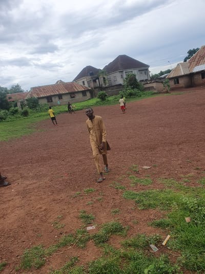 This image, captured in Idah, Nigeria, depicts an outdoor scene on a reddish-brown dirt field with interspersed patches of green grass. Four individuals are present, engaging in recreational activities, against a backdrop of various residential structures under an overcast sky.
In the foreground, slightly to the right of the center, a dark-skinned male subject is walking towards the viewer, smiling directly at the camera. He wears a light brown or tan long-sleeved tunic and matching trousers, with light blue sandals on his feet. His left hand is partially visible, holding a dark, rectangular object against his thigh.
In the mid-ground, three additional male subjects are visible. To the left, a subject in a yellow t-shirt and dark shorts stands facing slightly away from the camera. In the center-right, a subject in a dark t-shirt and dark shorts is actively engaged with a black and white football (soccer ball) at his feet, suggesting play. Further to the right and slightly behind, a fourth subject in a red top and dark shorts is also on the field, partially obscured, and appears to be involved in the same activity. A makeshift net, possibly for volleyball or football, is stretched horizontally across the mid-background between two unidentified supports.
The ground consists of reddish-brown laterite soil, characteristic of the region, with numerous small pebbles and debris. Scattered clumps of green grass grow unevenly across the field. In the background, a series of buildings are visible. To the left, a single-story structure with a rusty corrugated metal roof and unpainted concrete block walls features multiple windows. In the center-background, a larger, two-story grey building with a dark-colored hip roof and white-framed windows is prominent. A water tank is mounted on a raised platform to the right of this building. Another structure with a rusty corrugated roof and unpainted walls is visible further to the right. Lush green trees and foliage are present throughout the background, situated behind and between the buildings. The sky is uniformly overcast with grey clouds. Litter, including a yellow wrapper and small white boxes, is visible on the ground in the foreground-right.