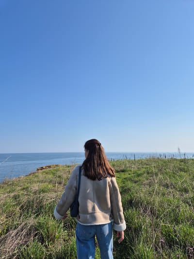 A woman stands on a grassy verge overlooking the Black Sea in Sveti Nikola, Bulgaria. She is wearing a beige shearling jacket, blue jeans, and has long brown hair. The sky is clear and blue. The scene is primarily composed of the sky, the sea, and the grassy verge.