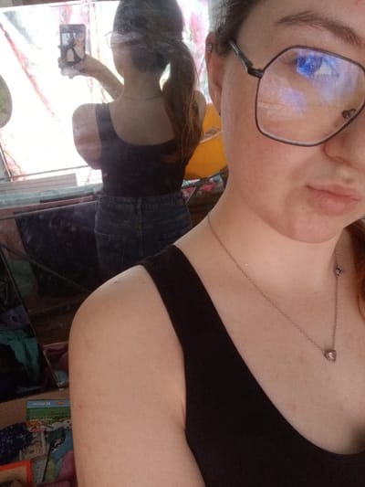 A person is depicted taking a mirror selfie within an indoor setting in Старый Шагирт, Russia. The foreground shows the right side of the individual's face, shoulder, and upper chest. The person wears large, dark-framed, geometric glasses, with blue reflections visible in the lenses. A thin silver-colored necklace with a small pink heart-shaped pendant rests on the chest. The reflection in the mirror shows the individual from the back, holding a black smartphone with both hands to capture the image. The person in the reflection wears a black tank top and light blue denim jeans, with long brown hair tied in a ponytail. The mirrored background reveals a cluttered room with a light-colored curtain displaying faint patterns, a horizontal surface, and various objects including folded clothing, a cardboard box, and a prominent bright yellow item.