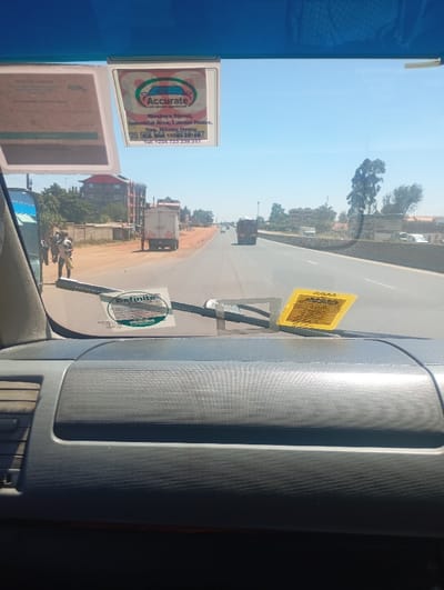 The image presents a forward-facing view from inside a vehicle onto a multi-lane highway in Muguga ward, Kenya, under a clear blue sky. The vehicle's dark grey, textured dashboard occupies the lower foreground, with two windshield wipers visible near the base of the windshield. Multiple stickers are affixed to the windshield: a partially obscured light-colored rectangular sticker is top-left, a prominent white rectangular sticker for "Accurate auto glass specialists Ltd" (Blashara Street, Industrial Area, Nairobi) is top-center, a green circular "Definite" sticker is bottom-left, and a yellow rectangular sticker is bottom-right.

On the left side of the highway, a dirt shoulder is visible. Approximately three to four individuals stand near a parked or stopped white box truck. A blue vehicle is partially obscured further left. Beyond the shoulder, a fence precedes a multi-story building with a red roof, accompanied by smaller structures and sparse vegetation. The asphalt highway features white lane markings and extends into the distance. A concrete median barrier separates the lanes on the right. Several vehicles, including trucks, are visible traveling down the road. Scattered trees and bushes line the roadside and horizon under sunny conditions.
