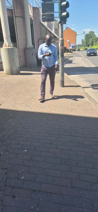 A man is walking on a brick sidewalk next to a street in Mainz, Germany. He is wearing a light blue long-sleeved shirt and dark pants. He is holding a smartphone in his right hand and looking down at it. The sidewalk is in shadow, cast by a pedestrian overpass. Behind the man, there is a traffic light, a street with cars, and a building. The sky is clear and blue.