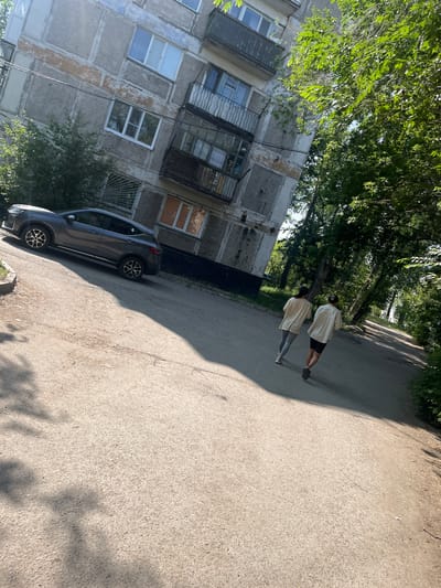The image shows a residential scene in Aksu, Kazakhstan. Two individuals are performing push-ups on a paved surface, which appears to be a road or sidewalk. They are dressed casually, with one person wearing a light-colored t-shirt and dark shorts, and the other wearing a similar light-colored t-shirt and grey leggings.
To the left of the individuals is a multi-story apartment building. The building has a weathered facade, with visible signs of age and disrepair. It features numerous windows, some with visible bars or grates. Trees with lush green foliage are present, casting shadows on the ground and the building, suggesting a time of day when the sun is casting long shadows, likely late afternoon or early morning. The overall atmosphere is one of a normal day in a residential area.
A dark-colored car is parked on the paved surface near the apartment building. The car is a modern compact SUV, and its presence indicates everyday life in the location. The ground is a textured asphalt, typical of urban or suburban streets. The sky is not directly visible due to the angle of the shot and the canopy of trees. There is no visible text in the image. The scene suggests an outdoor activity taking place in a residential neighborhood.