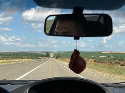 The image shows a view from the inside of a car driving on a highway in Solenyi, Russia. The driver's reflection is visible in the rearview mirror. In the distance, a flat landscape with fields and some buildings can be seen. A red, fuzzy air freshener hangs from the rearview mirror. The sky is blue with scattered clouds.
