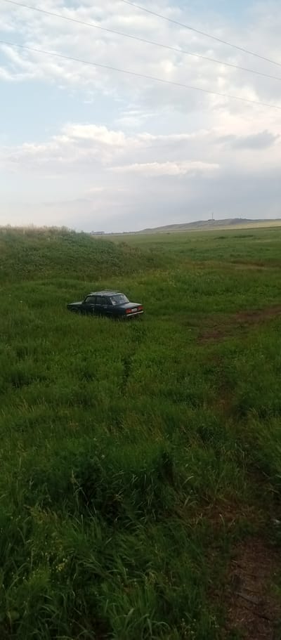 A dark-colored sedan, possibly a Lada, with a roof rack, is parked in a lush, green grassy field. The car is the primary object, positioned slightly off-center to the left. The setting is a wide-open, rural landscape with rolling hills in the distance. A dirt path is visible in the lower right corner of the image. In the background, a tall tower or antenna structure stands on one of the distant hills. The sky is partly cloudy, with soft, diffused light suggesting it might be late afternoon or early evening. The overall impression is one of solitude and stillness, with no people visible. The location is identified as Marinovka, Kazakhstan. No discernible text is visible on the car or in the surrounding environment.
