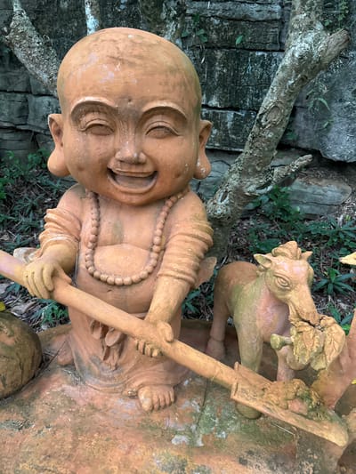 A terra cotta statue of a cheerful, bald-headed child with large ears and a wide grin is the central subject. The child is wearing a beaded necklace and appears to be holding a wooden staff with both hands. To the right of the child is a smaller terra cotta statue of a horse. The statues are situated outdoors, with a stone wall and trees in the background. The ground is covered with dry leaves and some greenery. This statue is located in the historical city of Tây Hoa Lư Ward, Vietnam.