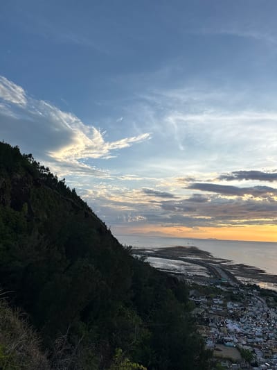 The image depicts a dramatic sunset over the sea, viewed from a densely forested hillside. The sky is a canvas of blues and oranges, with streaks of white and grey clouds catching the fading sunlight. The sun, low on the horizon, casts a warm, golden glow across the water, which appears calm and reflective.
In the distance, along the coastline, a cluster of buildings is visible, possibly a village or town, nestled against the water's edge. The architecture suggests a coastal community. The sea itself shows signs of low tide, with exposed mudflats or sandbanks reflecting the sky's colors.
The foreground is dominated by the dark, silhouetted mass of a hillside covered in thick vegetation. The trees and bushes are rendered in deep green and black, creating a stark contrast with the bright sky and sea. The overall impression is one of natural beauty and tranquility, with the human settlement appearing small and integrated into the grander landscape.
The time of day is clearly sunset, indicated by the warm, low-angle light and the colors in the sky. The weather appears clear, with the clouds contributing to the visual spectacle of the sunset rather than obscuring it. The location cues, such as the coastal settlement and the typography of the hillside, are consistent with a Vietnamese coastal setting. There are no visible people or specific objects of interest that draw attention. The scene evokes a sense of peaceful contemplation as day transitions into night.