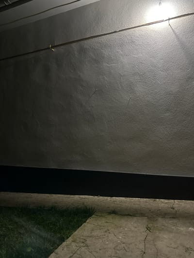 This image shows a close-up view of a textured wall, likely made of concrete or stucco, painted a dark grey. A bright light source is visible on the left side, casting a strong glare and illuminating a vertical wire or cable running down the wall. There are a few small, light-colored objects, possibly nails or imperfections, embedded in the wall's surface.
To the right of the grey wall, there is a narrow vertical strip of black, which could be a shadow or a separate architectural element. Further to the right, another wall or surface is visible, this one lighter in color and with a rough, cracked texture. This surface is partially covered in green grass and dry, thin vegetation, suggesting it might be an exterior wall or boundary where plants are growing. The lighting suggests it is either nighttime or a very dimly lit indoor space, with the prominent light source creating strong contrasts. The overall impression is one of an outdoor or semi-outdoor utilitarian space. No people, text, or specific events are depicted.