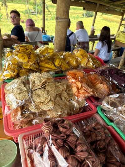 In Penebel, Indonesia, this image captures a market stall at a rice terrace overlook. In the foreground, several plastic-wrapped bags of snacks are displayed in red and green baskets. The snacks include yellow banana chips, flat, circular chips, ridged orange chips, and dark purple chips, along with some dark brown crisps. In the background, four people are seated at a wooden table, enjoying the view of the lush green rice terraces. A man in a black shirt and glasses is on the left, facing away from the camera, while a woman with pink hair sits beside him. Two other individuals, a woman in a white shirt and a woman in a white t-shirt, are also seated. The stall is constructed with bamboo, providing a rustic and natural ambiance fitting for its scenic location.