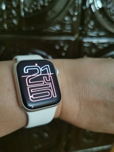 A close-up shot shows a person's wrist wearing a white-banded smartwatch with a black screen displaying the time "21:10" in a stylized, neon-pink font. The background is dark and out of focus, with some blurry highlights suggesting a textured surface or decorative element. The image is shot from a slightly elevated angle, focusing on the watch face and the skin of the wrist. The Pociumbeni, Moldova location is not discernible from the image.