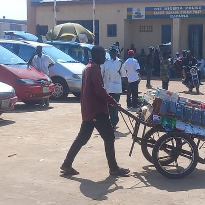 A man in a dark red shirt and dark pants pushes a two-wheeled cart loaded with cell phone accessories on a dirt road in front of a police station. The cart is filled with various items, including cell phone cases and boxes. Behind the cart, several people are visible, some standing and some sitting. A motorcycle is parked to the right of the frame. The building in the background has a sign that reads "THE NIGERIA POLICE LAGOS GARAGE POLICE POST KADUNA." The scene appears to be a busy street or market area in Hayin Banki, Nigeria.